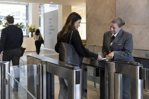 Illustrative private security work in Hartford, Connecticut: Mature woman security officer in grey security blazer at an insurance office turnstile helping an employee scan their access badge.