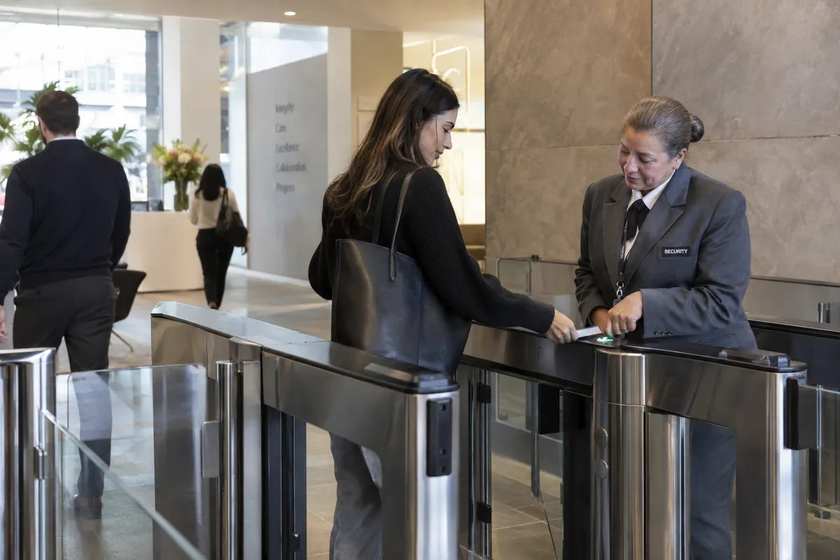 Illustrative private security work in Hartford, Connecticut: Mature woman security officer in grey security blazer at an insurance office turnstile helping an employee scan their access badge.