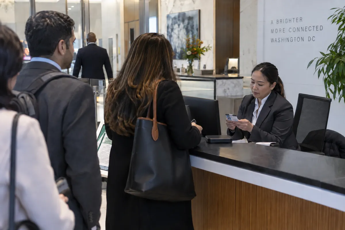 Illustrative private security work in Washington, District of Columbia: Asian female private security officer in a charcoal suit managing visitor screening at an ordinary private office lobby reception desk in Washington DC.