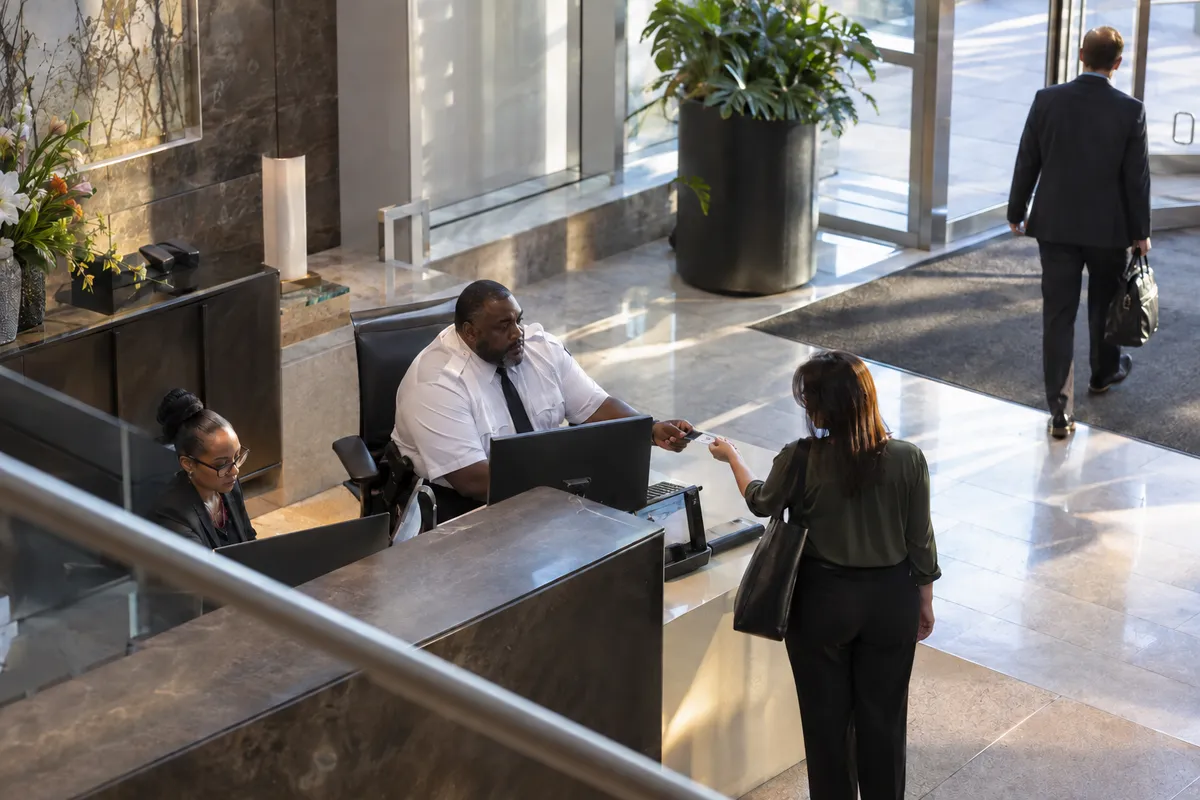 Illustrative private security work in Atlanta, Georgia: Black male private security officer in white uniform shirt and navy tie working a busy modern office-lobby access desk in Atlanta, visitors arriving.