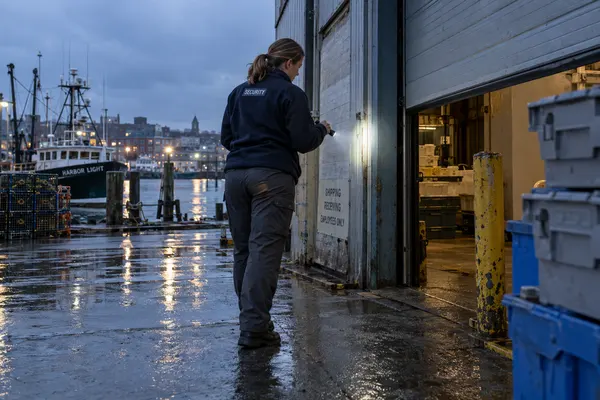 Illustrative private security work in Portland, Maine: Female private security officer in navy fleece and grey work trousers inspecting a harbour warehouse entrance with a small flashlight after rain, working waterfront in Portland Maine.