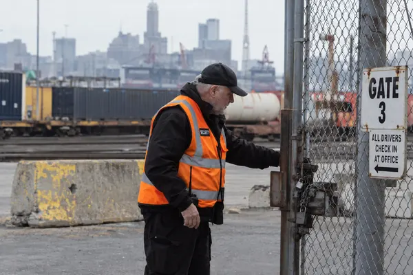 Illustrative private security work in Baltimore, Maryland: Older male private security officer in high-vis orange vest over black work jacket checking an industrial gate at a Baltimore freight facility.