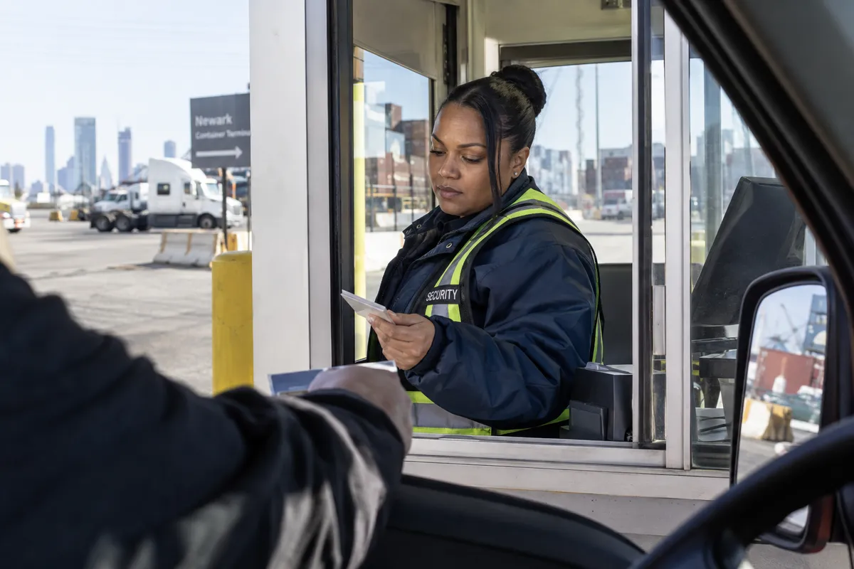 Illustrative private security work in Newark, New Jersey: Female private security officer in navy windbreaker and reflective vest checking shipping access from a gate booth in Newark New Jersey, glancing at driver's pass.