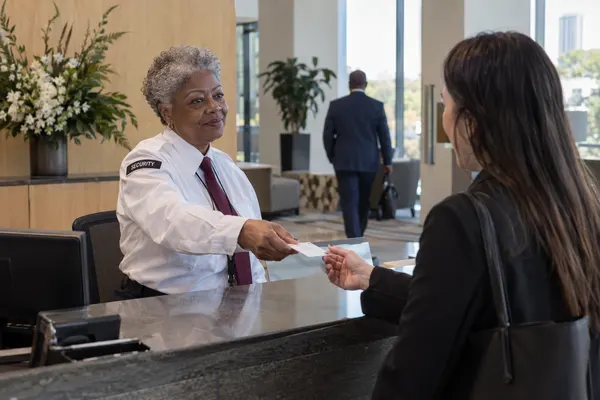 Illustrative private security work in Richmond, Virginia: Older female private security officer in white security shirt and burgundy tie handing a visitor pass to a arriving office visitor at corporate reception in Richmond Virginia.