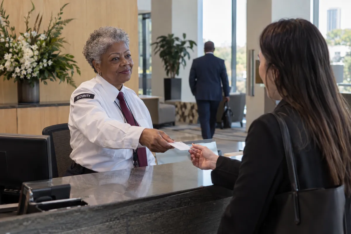 Illustrative private security work in Richmond, Virginia: Older female private security officer in white security shirt and burgundy tie handing a visitor pass to a arriving office visitor at corporate reception in Richmond Virginia.